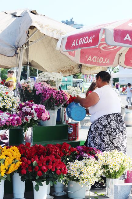 A floral display at a street market featuring a variety of fresh flowers arranged in white and green buckets, including vibrant red roses, bright yellow sunflowers, and clusters of white and pink chry
