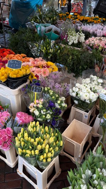 A vibrant display of fresh flowers arranged in multiple bouquets and stems at a flower market stall. There are yellow tulips in a white crate at the front, alongside pink roses and purple lisianthus. 
