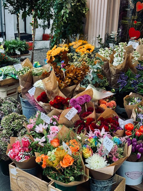 A display of freshly arranged flower bouquets at a florist shop, featuring a variety of blooms wrapped in brown craft paper and some in clear glass vases. The arrangement includes sunflowers, roses, l