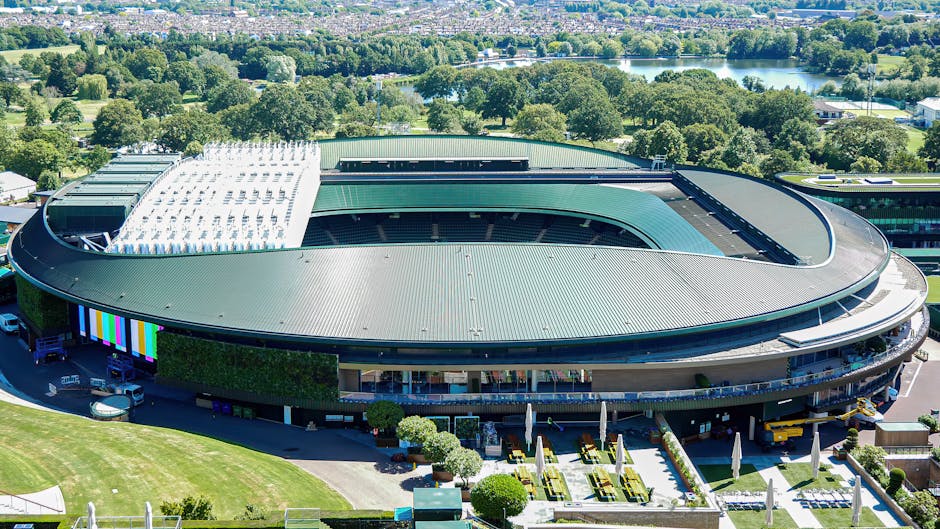 An aerial view of the All England Lawn Tennis Club featuring a large, oval-shaped building with a green metal roof and surrounding lush greenery, situated near a river. In the foreground, there are ne
