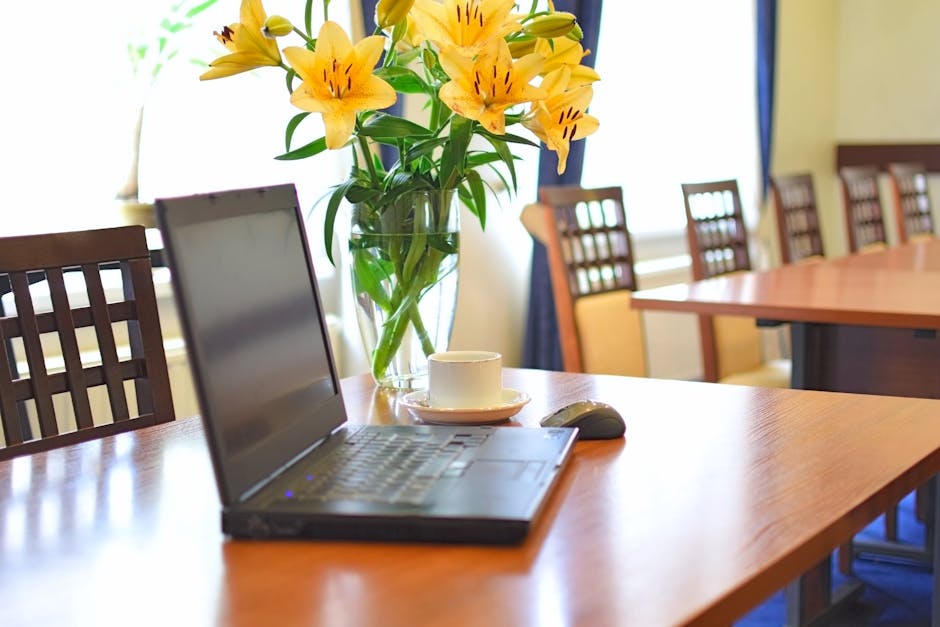 A clear glass vase filled with vibrant yellow lilies and green foliage, placed on a polished wooden conference table. The lilies have open petals with dark speckles in the center, showcasing their fre