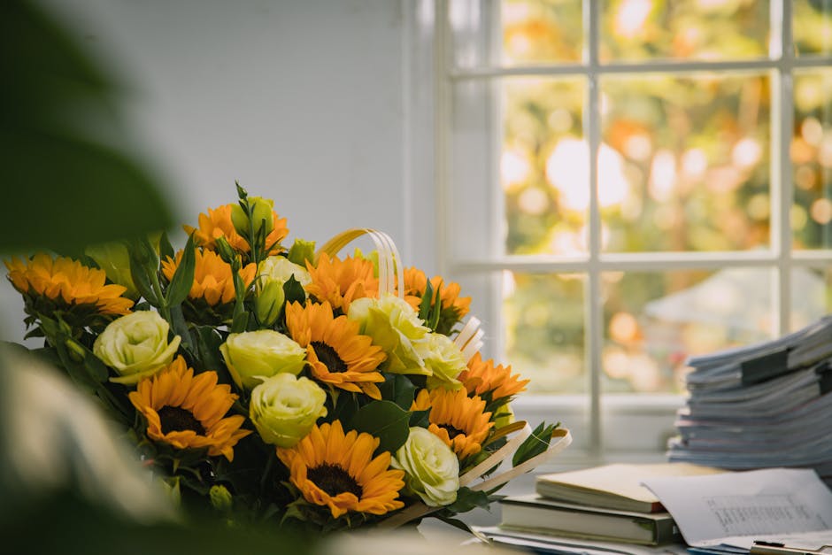 A vibrant bouquet of fresh flowers featuring bright yellow sunflowers, pale yellow lisianthus, and green foliage arranged in a round style. The bouquet is placed on a desk near a large window with nat