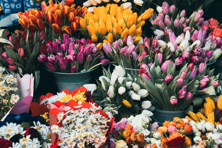 A vibrant display of fresh flowers including tulips in shades of pink, white, orange, and purple, arranged in metal buckets and floral bouquets. The tulips have long green stems and petals with a smoo