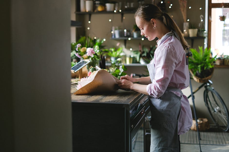 A female florist in a light pink shirt and gray apron arranging a bouquet of fresh flowers on a wooden countertop in a cozy, well-lit flower shop. The bouquet is wrapped in brown craft paper, featurin
