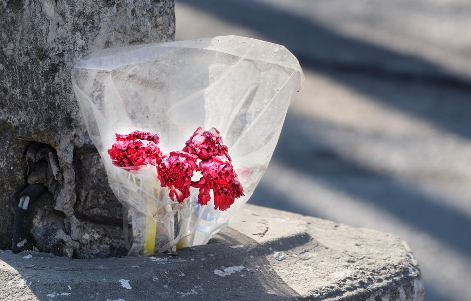 A small bouquet of wilted pink carnations wrapped in clear cellophane rests against a weathered stone wall on a concrete ledge. The carnations show signs of fading and petal browning, indicating they 