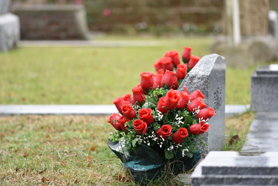 A bouquet of fresh red roses with lush green foliage and small white filler flowers, arranged around the stems and wrapped in dark green and black floral paper, is placed upright against a grey granit